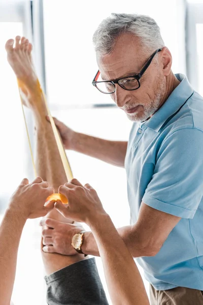 Médecin dans des lunettes debout près de l'homme faisant de l'exercice avec des sangles de suspension pendant la réadaptation — Photo de stock