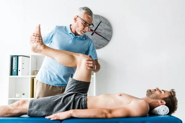 Guapo paciente acostado en la mesa de masaje y haciendo ejercicio cerca del médico en gafas - foto de stock