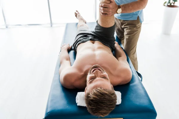 Vista recortada del médico cerca del paciente acostado en la mesa de masaje y haciendo ejercicio en la clínica - foto de stock