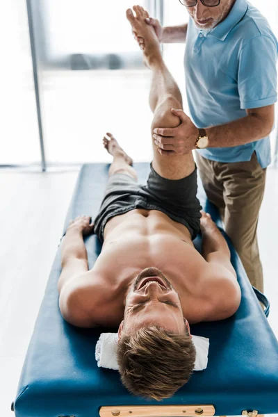 Vista recortada del médico cerca del paciente con los ojos cerrados acostados en la mesa de masaje y haciendo ejercicio en la clínica - foto de stock