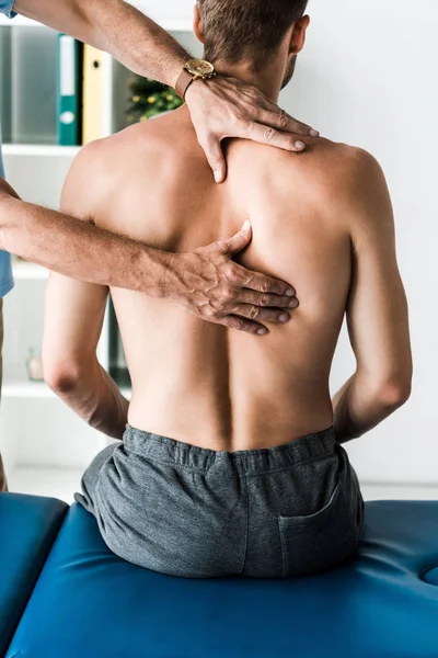 Vista recortada del médico tocando la espalda del paciente sin camisa sentado en la mesa de masaje - foto de stock