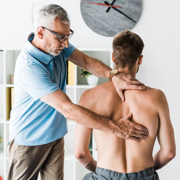 Médecin barbu dans des lunettes touchant le dos d'un patient torse nu à la clinique — Photo de stock