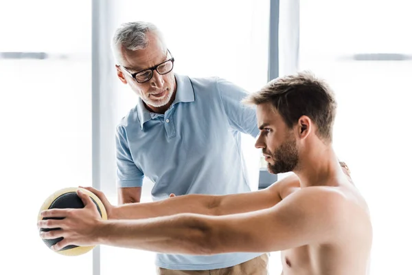 Bearded doctor in glasses looking at patient working out with ball — Stock Photo