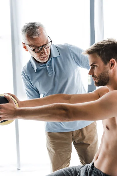 Enfoque selectivo del médico barbudo en gafas mirando al paciente sin camisa haciendo ejercicio con pelota - foto de stock