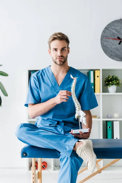 Médico bonito olhando para a câmera enquanto segurando modelo de coluna na clínica — Fotografia de Stock