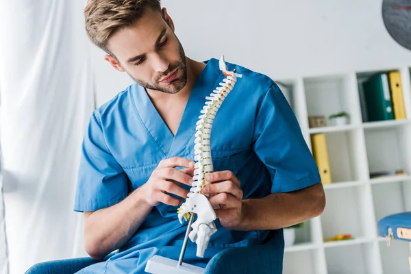 Medico barbuto toccando modello di colonna vertebrale mentre seduto in poltrona — Foto stock