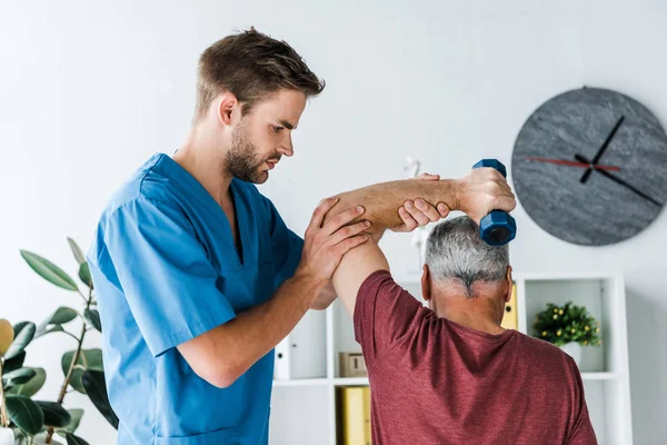 Вид сзади мужчины средних лет, выполняющего упражнения с гантелями возле врача — стоковое фото