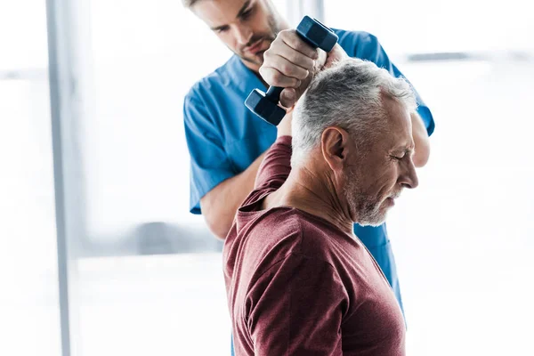 Enfoque selectivo del hombre de mediana edad haciendo ejercicio con mancuerna cerca del médico - foto de stock