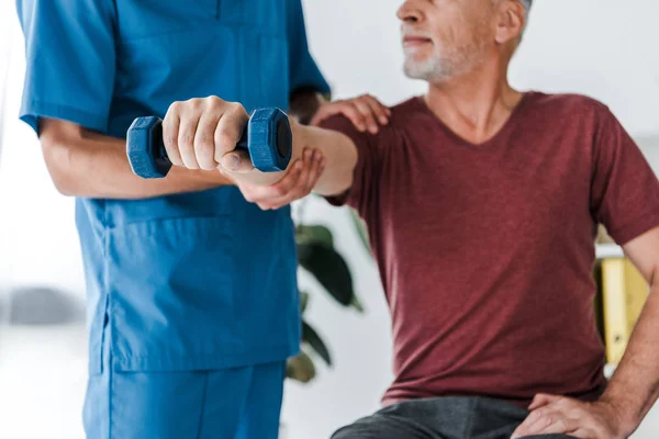 Vue recadrée du médecin debout près d'un patient d'âge moyen faisant de l'exercice avec haltère — Photo de stock