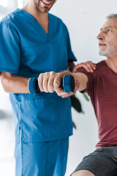 Обрезанный вид счастливого врача, стоящего рядом со зрелым мужчиной, упражняющимся с гантелями в клинике — стоковое фото