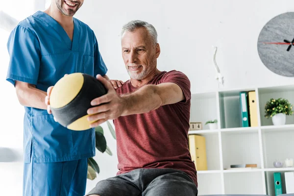 Vista recortada de médico feliz cerca de hombre maduro haciendo ejercicio con pelota - foto de stock