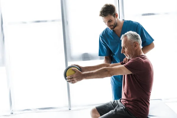 Hombre de mediana edad haciendo ejercicio con pelota cerca de médico guapo - foto de stock