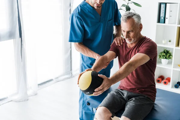 Cropped view of doctor standing near mature man exercising with ball — Stock Photo