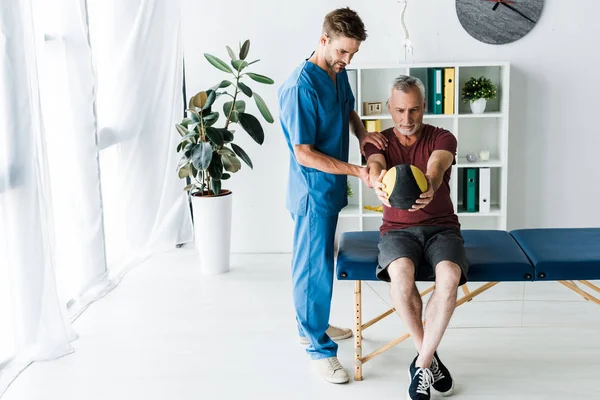 Handsome doctor standing near mature man working out with ball — Stock Photo