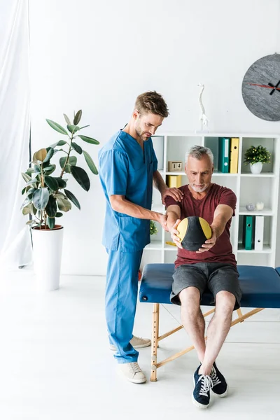 Handsome doctor standing near mature man exercising with ball — Stock Photo