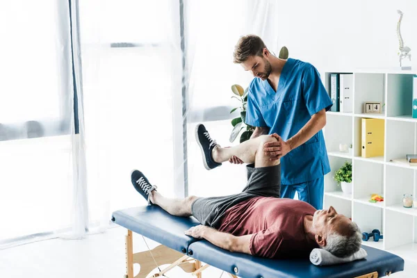 Schöner Arzt berührt Bein eines reifen Patienten liegend auf Massagetisch — Stockfoto