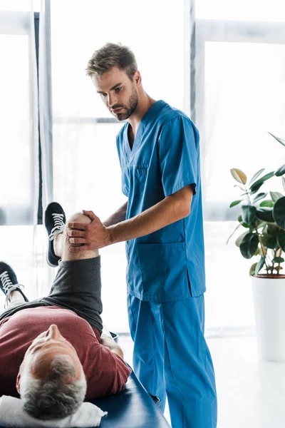Médico bonito tocando perna de paciente maduro exercitando na mesa de massagem — Fotografia de Stock