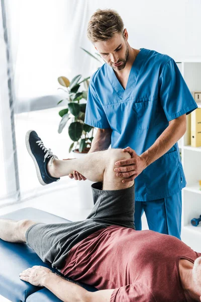 Médico barbudo tocando perna de paciente maduro exercitando na mesa de massagem — Fotografia de Stock