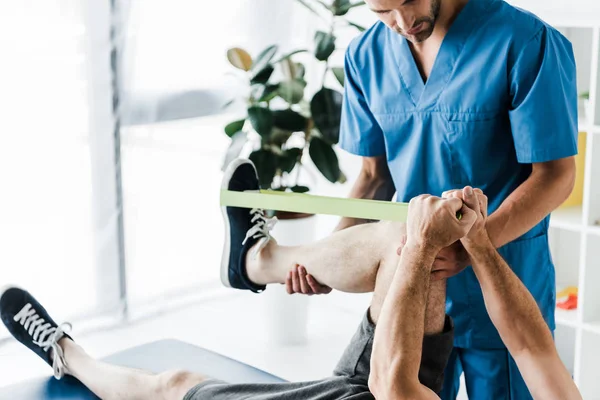 Vista ritagliata del medico che tiene la gamba sull'uomo maturo formazione con cinghie di sospensione — Foto stock