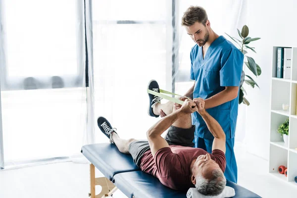 Guapo doctor barbudo sosteniendo pierna de paciente maduro haciendo ejercicio con elásticos - foto de stock