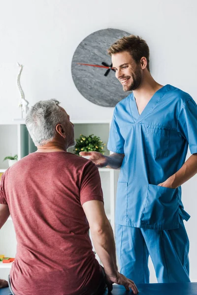 Glücklicher Arzt, der mit der Hand in der Tasche neben reifem Mann in der Klinik steht — Stockfoto