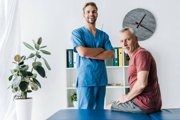 Handsome doctor standing with crossed arms near happy patient — Stock Photo