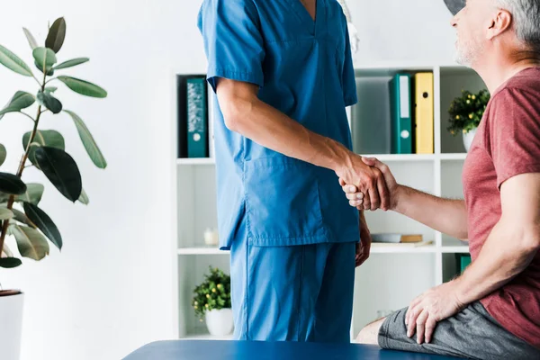 Обрезанный вид врача пожимающего руку с веселым зрелым пациентом в клинике — стоковое фото
