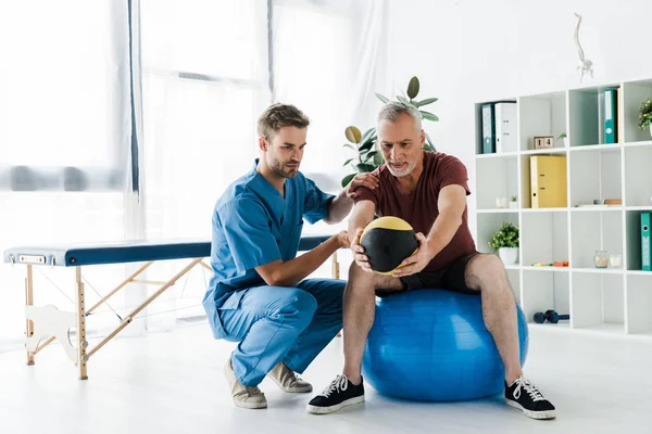 Reifer Patient trainiert auf Fitnessball in der Nähe des Arztes — Stockfoto