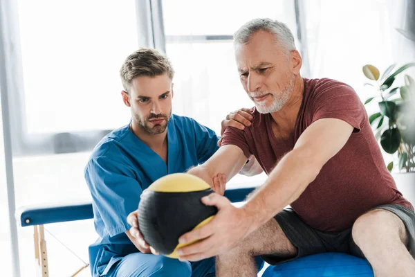 Médecin assis près d'un patient mature faisant de l'exercice sur balle de fitness — Photo de stock