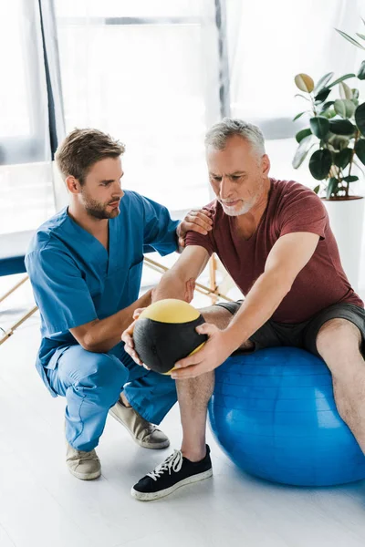 Красивый врач сидит рядом с зрелым пациентом упражнения на фитнес-мяч — стоковое фото