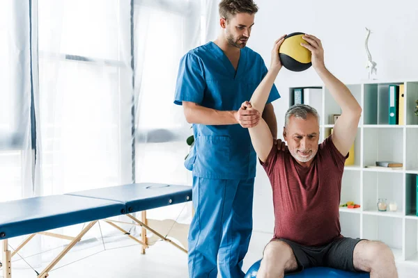 Красивый врач, стоящий рядом со зрелым мужчиной, тренирующимся на фитнес-мяч — стоковое фото