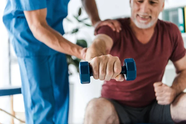 Selective focus of cheerful mature man working out with dumbbell near doctor — Stock Photo
