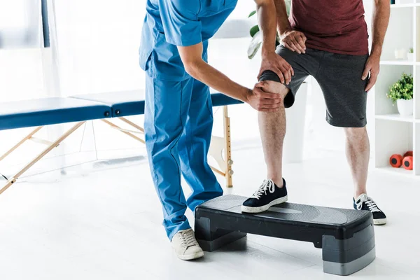 Обрезанный вид врача, трогающего колено взрослого пациента на ступенчатой платформе — стоковое фото