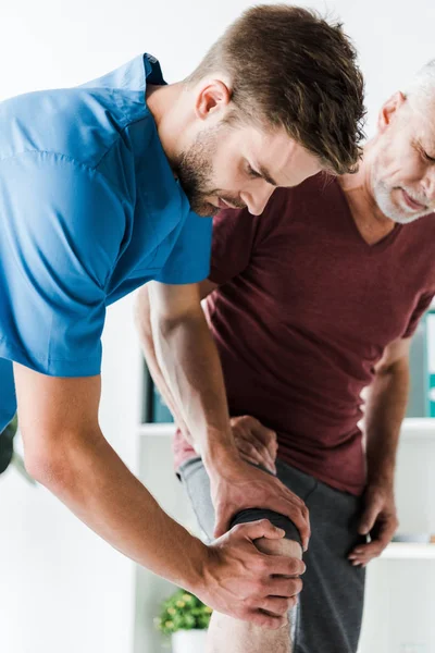 Handsome doctor touching knee of mature patient — Stock Photo