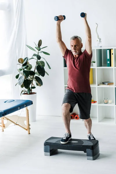 Middle aged man exercising with dumbbells on step platform in clinic — Stock Photo
