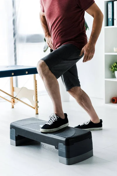 Vista recortada del hombre de mediana edad que hace ejercicio en la plataforma de paso en la clínica - foto de stock