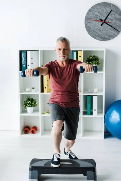 Barbuto uomo di mezza età che esercita con manubri sulla piattaforma passo in clinica — Foto stock