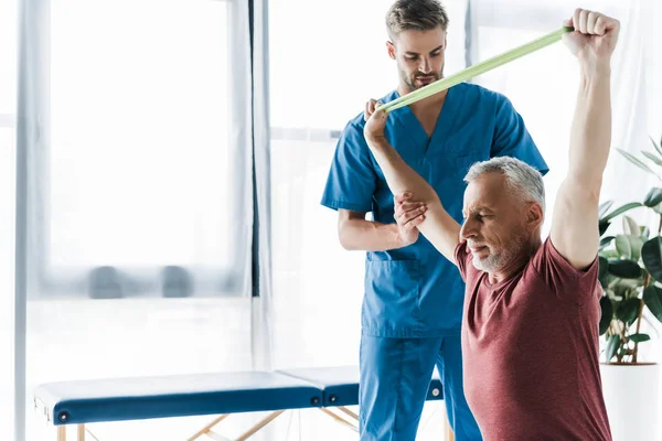 Arzt steht neben Mann mittleren Alters, der mit Widerstandsband trainiert — Stockfoto