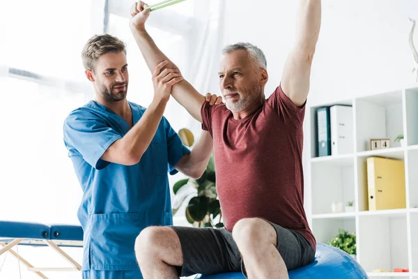 Médico cerca de hombre de mediana edad haciendo ejercicio con banda de resistencia - foto de stock