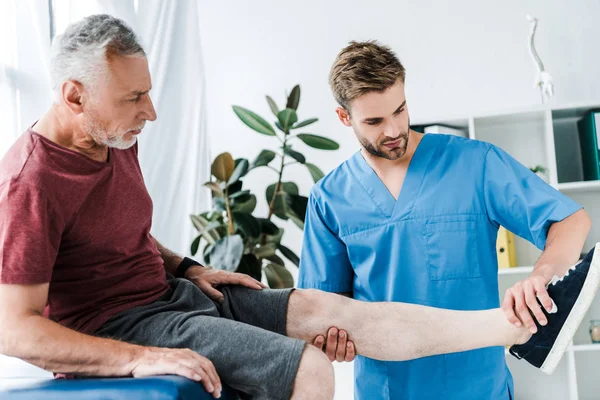 Handsome doctor holding leg on mature patient in clinic — Stock Photo