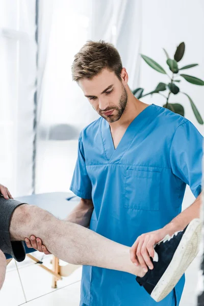 Guapo barbudo doctor sosteniendo pierna en maduro paciente en clínica - foto de stock