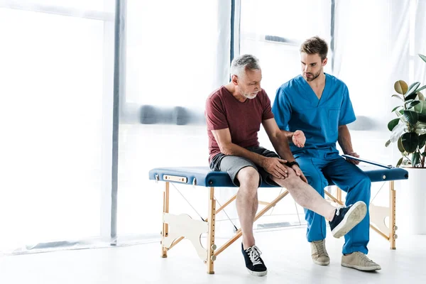 Bearded mature patient touching leg near doctor in clinic — Stock Photo