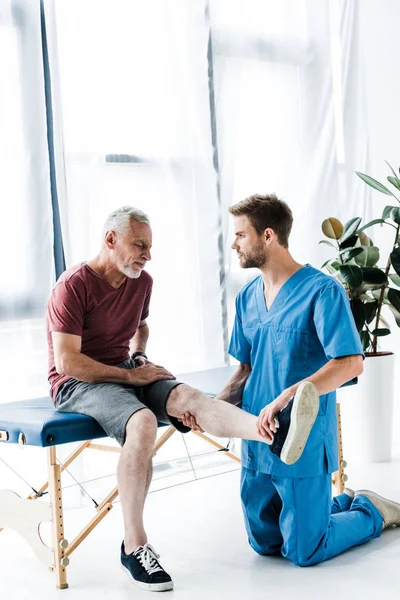 Schöner Arzt, der Bein berührt und bärtige reife Patientin ansieht — Stockfoto