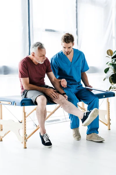 Bearded mature patient touching leg near doctor holding clipboard in clinic — Stock Photo