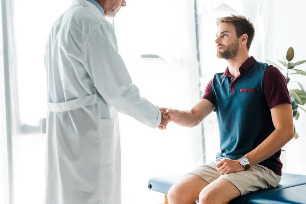 Cropped view of doctor in white coat shaking hands with handsome man — Stock Photo