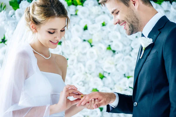 Atractiva y sonriente novia poniendo anillo de bodas en el dedo - foto de stock
