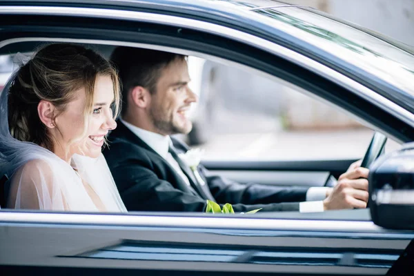 Foco seletivo de noiva atraente em véu de noiva e noivo sorrindo no carro — Fotografia de Stock