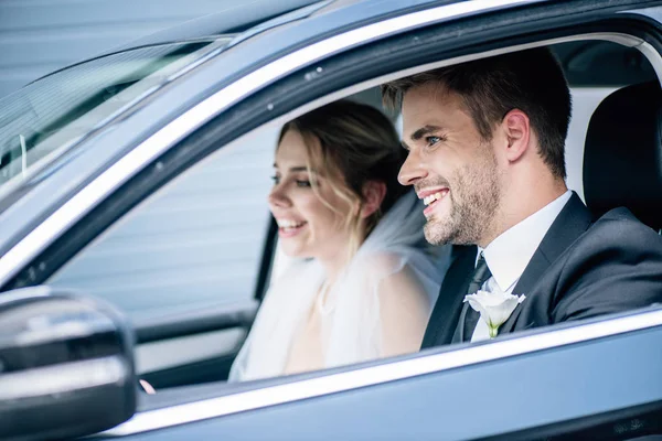 Selective focus of attractive bride in bridal veil and bridegroom smiling in car — Stock Photo