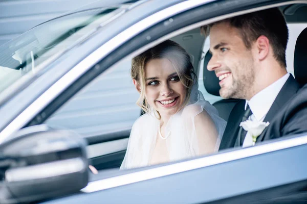 Noiva atraente em véu nupcial e noivo sorrindo no carro — Fotografia de Stock