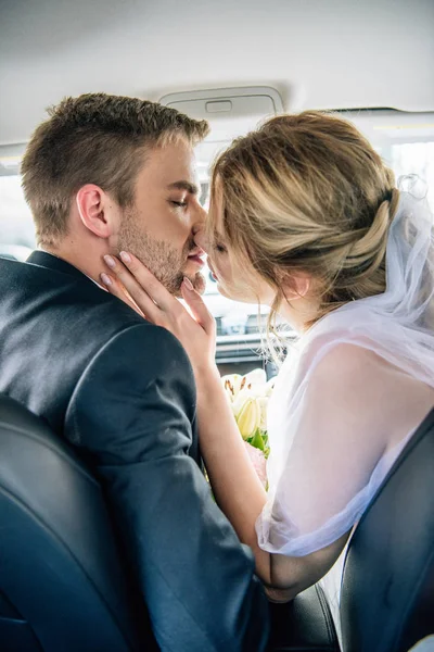 Back view of attractive bride in bridal veil and bridegroom kissing in car — Stock Photo